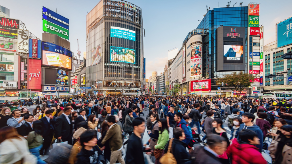 Busy Tokyo crosswalk in Japan, crowded with pedestrians under neon-adorned glass towers and bright digital billboards on a clear day.