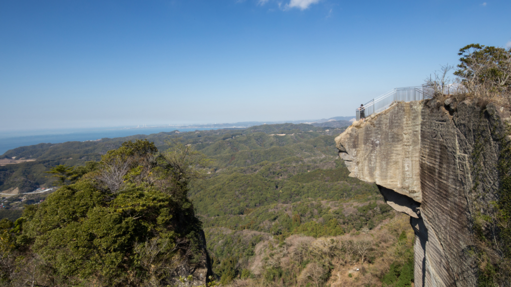 Dramatic cliff edge of Mt. Nokogiri with a metal railing and a person gazing over layered forested hills toward a distant coastline.