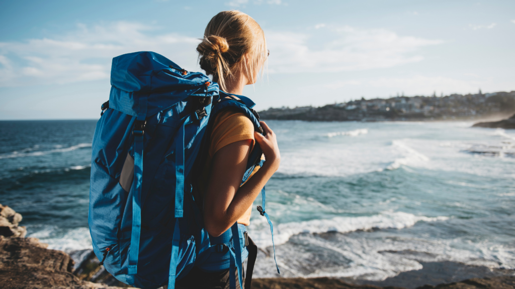Backpacker wearing a large blue hiking backpack gazes toward a rocky coastline, ocean waves rolling in under a bright sky.