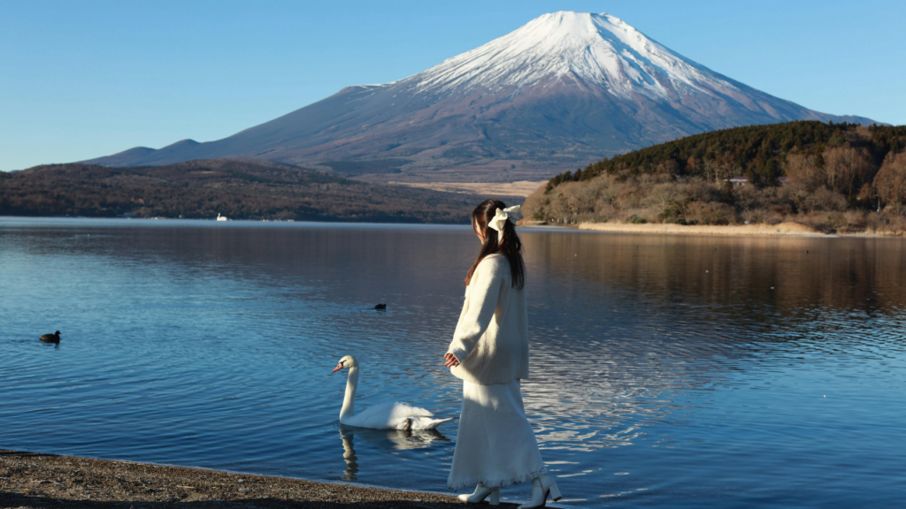 A woman in white walks along a lake as swans glide by, with snow-capped Mount Fuji near Tokyo, Japan.