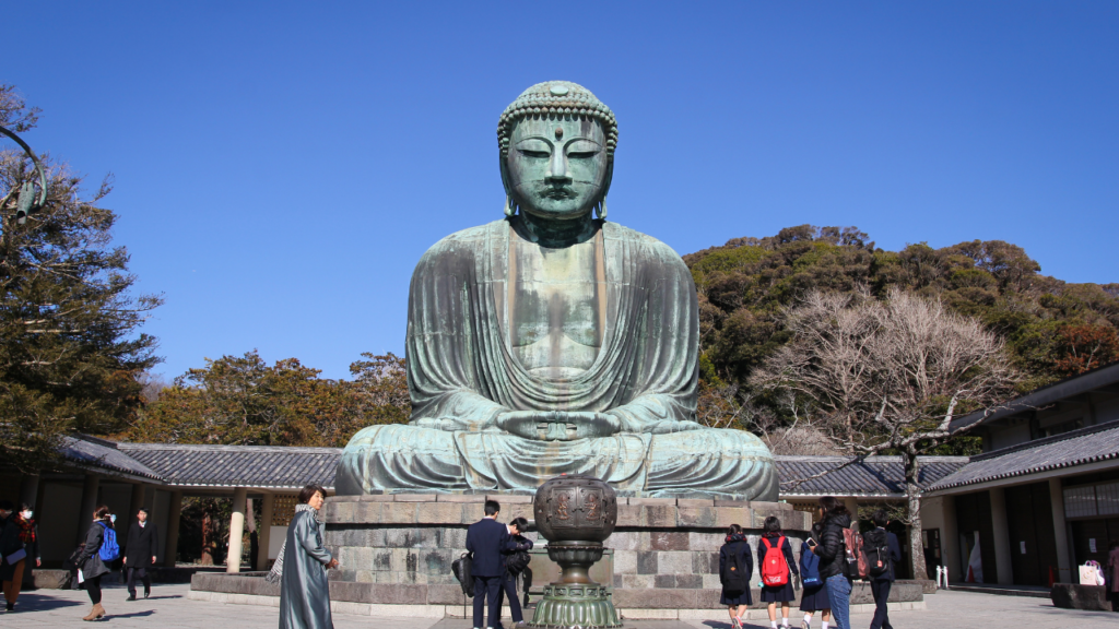 Massive bronze Buddha statue seated in meditation at a Japan temple courtyard, with visitors and tiled halls under a blue sky.