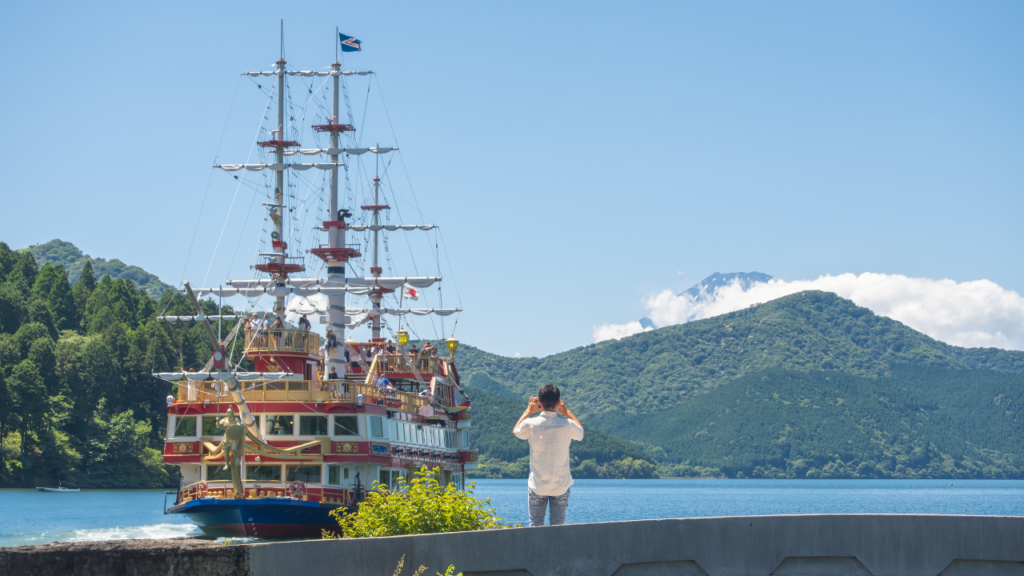 Colorful ornate ship with tall masts docked by a lake; a man takes a photo as green hills and distant Mount Fuji rise beyond.