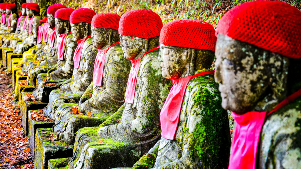 Line of mossy stone statues wearing red knit caps and pink bibs, along a leaf-strewn path in Japan.