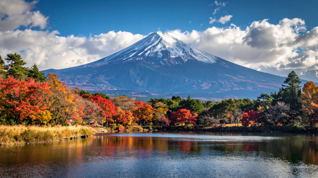 Snow-capped Mount Fuji rises over a vibrant autumn forest in Japan, reflected in a calm lake beneath blue sky and clouds.