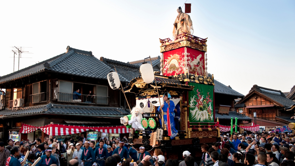 Elaborate festival float with gilded carvings and banners moves through a Kawagoe street, blue-robed performers aboard.