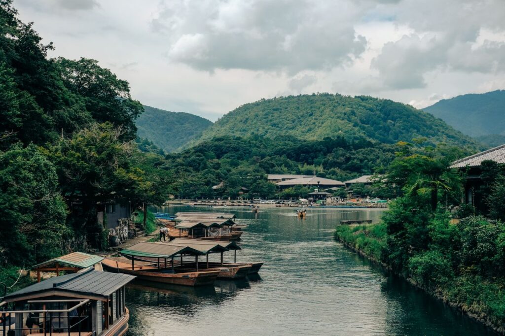 Calm river winding through a valley, in Kyoto with docked wooden boats along the shore and a distant village amid forested hills.
