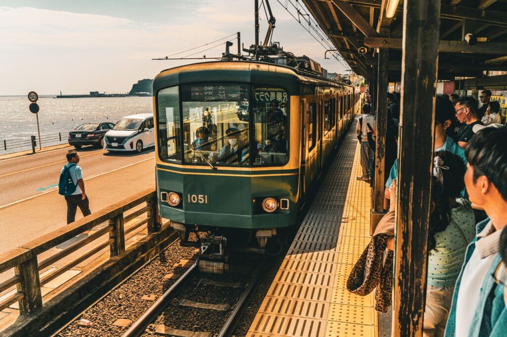 Teal-green tram at a seaside platform in Enoshima with passengers waiting under a shelter as the train glides past.