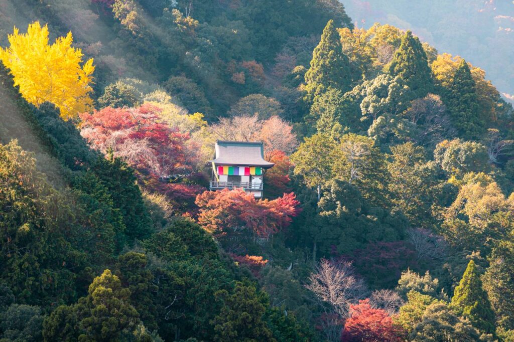 Traditional Japanese temple perched on a hillside balcony, amid vivid autumn foliage and colorful banners.