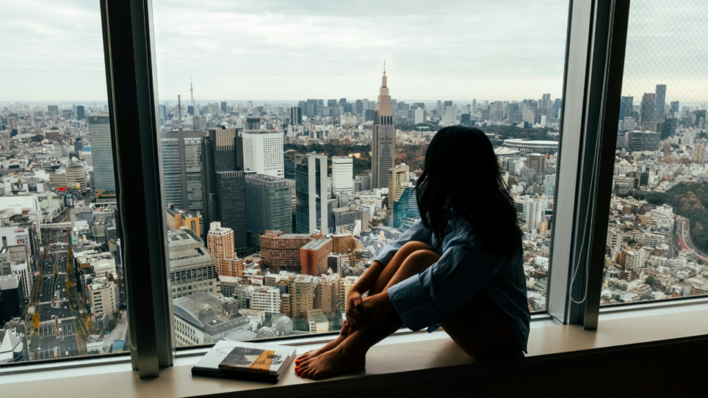 Woman sits barefoot on a window ledge, knees drawn up, overlooking a dense Tokyo skyline under an overcast gray sky, Japan.