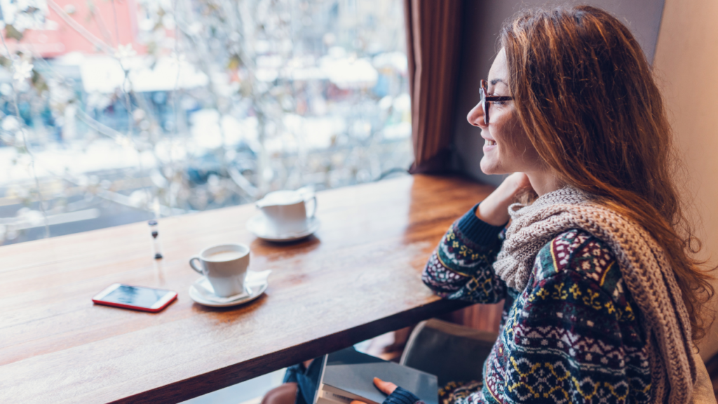 Smiling woman with glasses and a chunky scarf sits at a cafe counter by a window, coffee cups and a smartphone on the table.