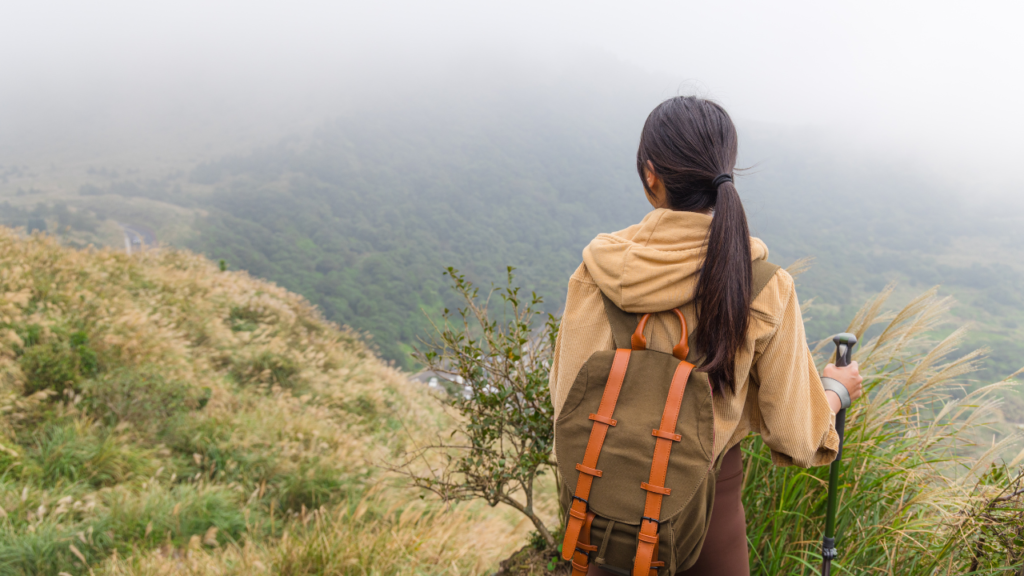 Hiker wearing a tan hoodie and olive backpack stands on a misty hillside, gripping a trekking pole, facing a foggy valley.