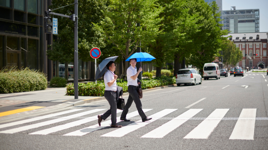 Two suited businessmen cross a crosswalk on a sunny Tokyo street in Japan, each with umbrella, blue or black, beside trees.