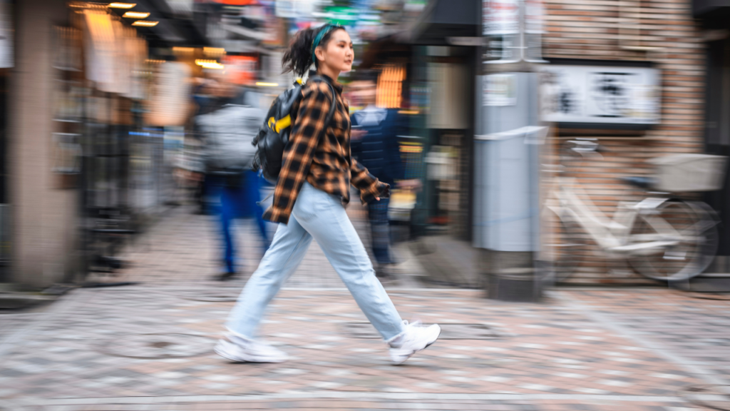Tokyo street scene with blur: a young woman in plaid shirt, light jeans and white sneakers walks past shops with a backpack.