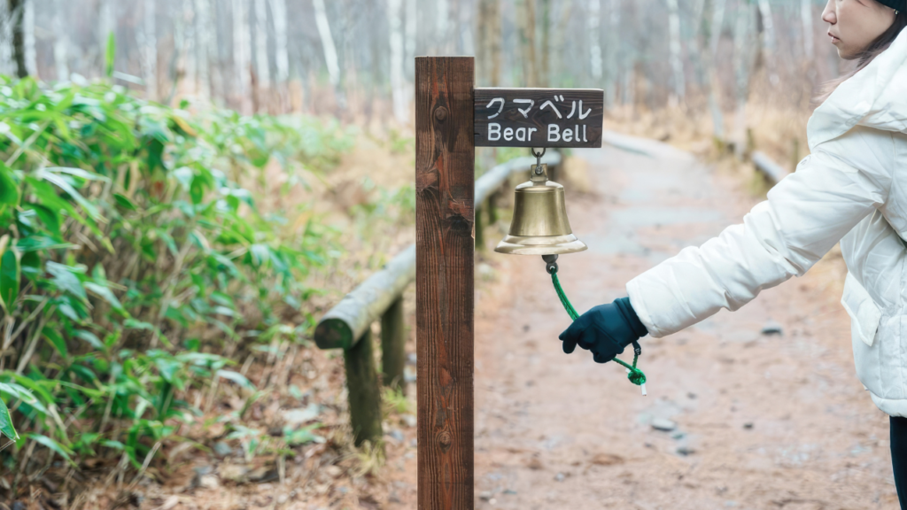 Person in a white jacket and dark gloves rings a bear bell on a wooden post beside a forest path in Japan.