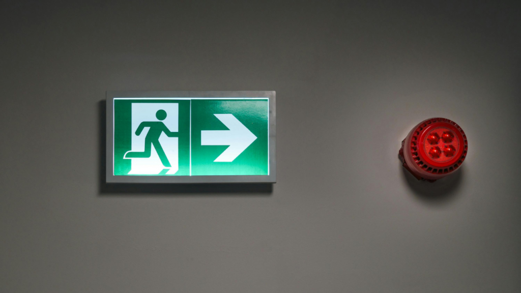 Green emergency exit sign with a running figure and right arrow, mounted on a plain wall beside a red fire alarm in Japan.