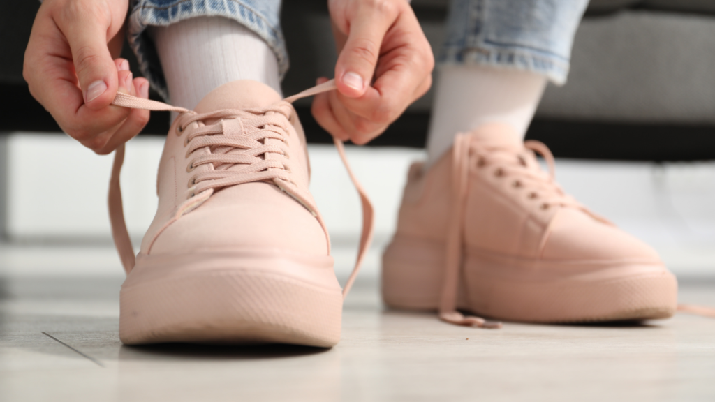 Close-up of pale pink sneakers being tied, hands threading laces on a tiled floor, worn with white socks and denim jeans, ready for a walk.