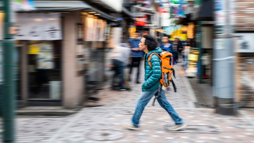 Motion-blurred Tokyo, Japan street scene with a man in teal puffer jacket and orange backpack crossing a cobblestone street.