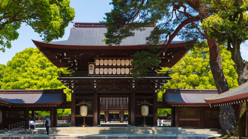 Traditional shrine gate at Meiji Shrine, Tokyo, Japan, framed by green trees as two visitors walk toward the inner courtyard.