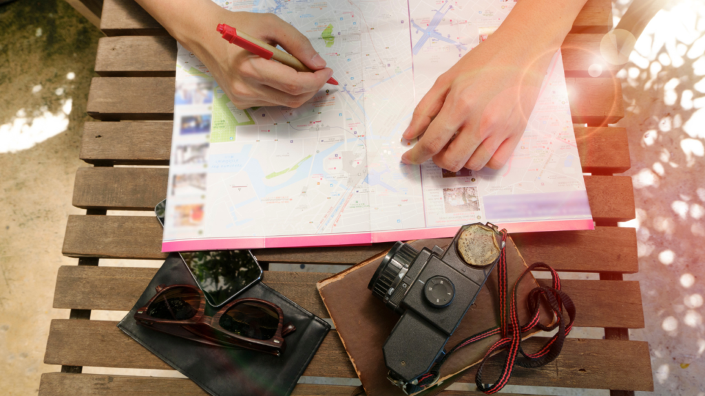 Two hands trace routes on a travel map spread on a wooden bench, beside a vintage camera, sunglasses, and a smartphone.
