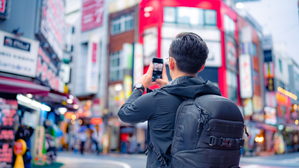 Back view of a traveler with a bulky black backpack snapping a photo on a busy Tokyo street lined with neon signs and shops.