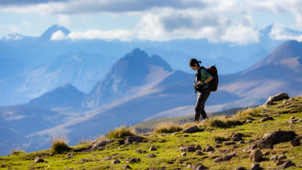 Hiker with a backpack traverses a grassy alpine ridge, rugged mountains fading into blue skies.